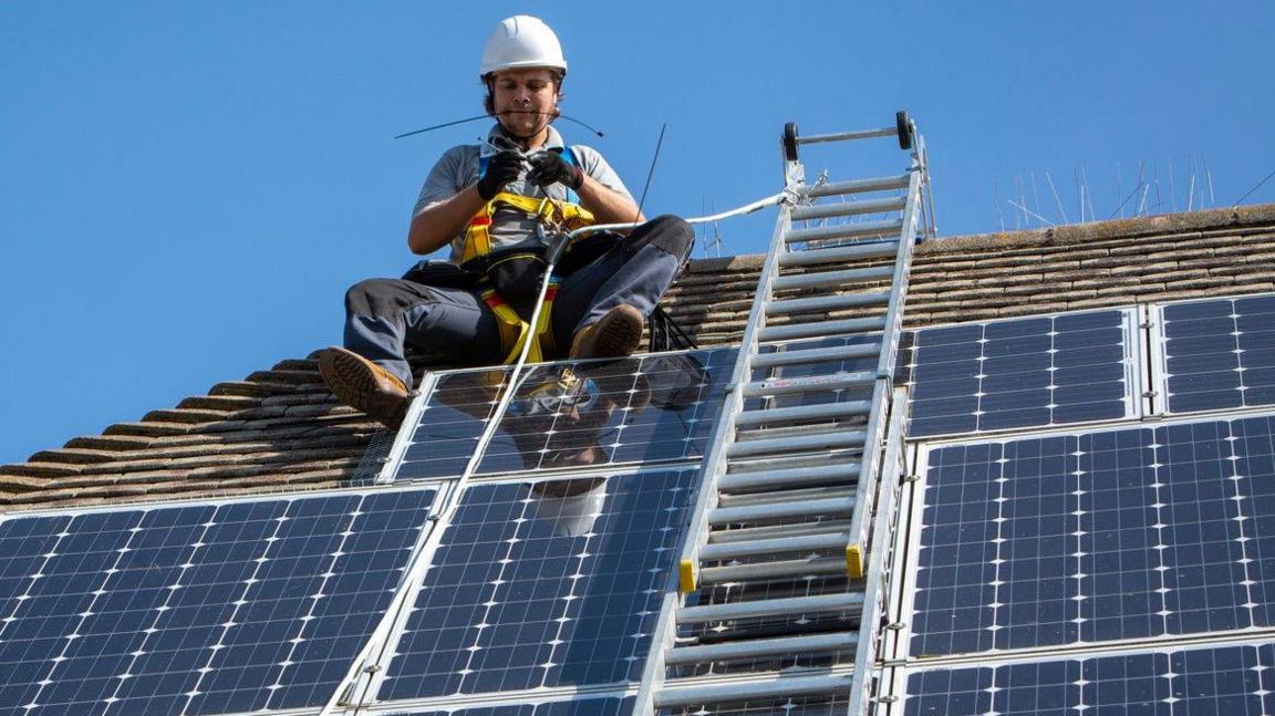 A maintenance engineer wearing a harness and hard hat sits on a roof next to a ladder, servicing solar panels around him on the roof. 