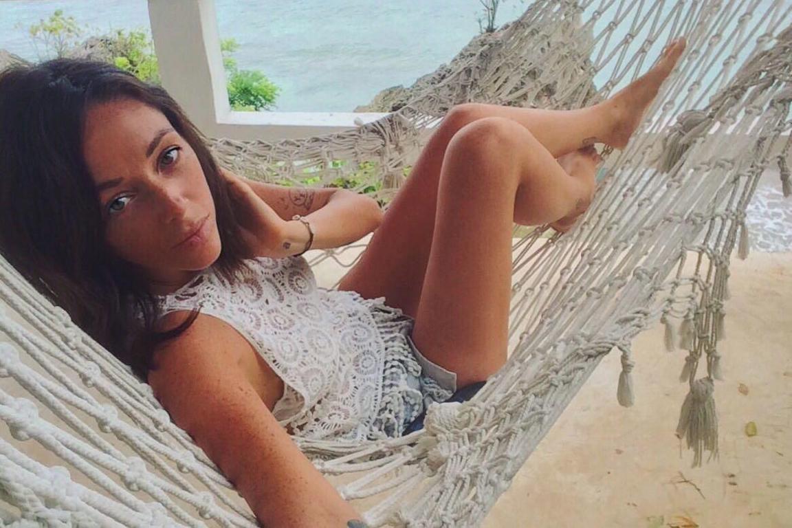 Kirsty McKie sits in a hammock and looks at the camera as she takes a selfie. The sea and a tree can be seen in the background and sand below her. She has brown hair that touches her shoulders.