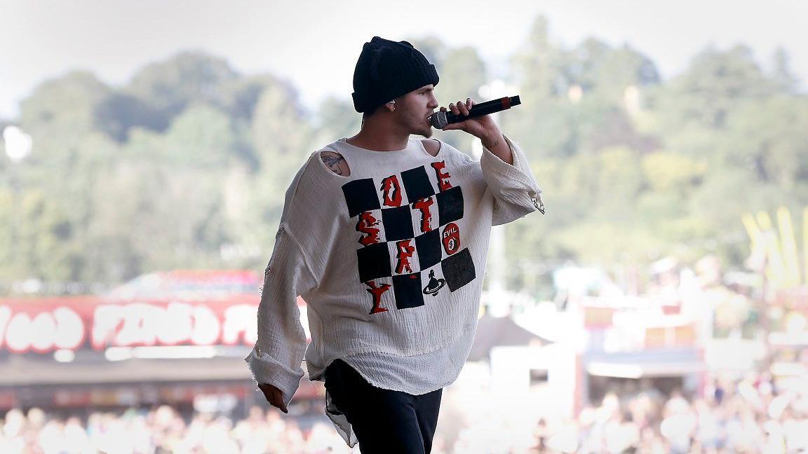 A man wearing a white top and a black hat singing into a microphone at a festival, with a crowd and some trees in soft focus behind him.