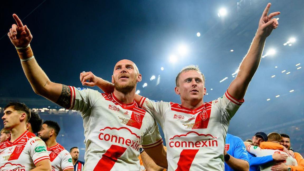 Hull Kingston Rovers players Dean Hadley (left) and Jez Litten (right) raise arms in celebration under the Old Trafford floodlights after beating Wigan Warriors in the Grand Final, with team-mates celebrating in the background.
