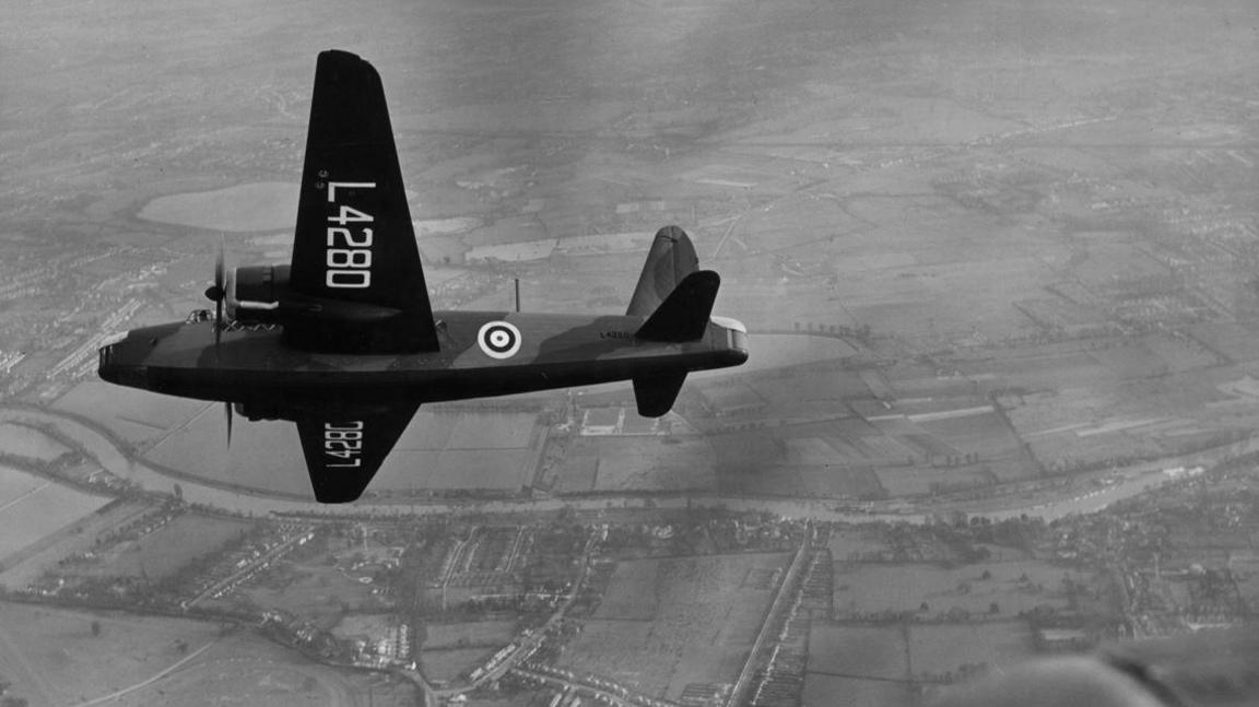 A black and white photo of a Wellington bomber flying over countryside. 