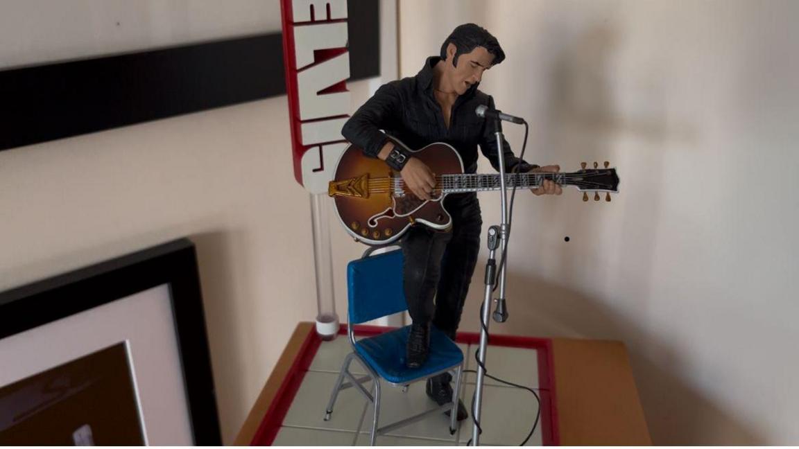 A 30cm (1ft) model of Elvis in black leather with his foot on a blue chair playing a sunburst electric guitar with microphone on a stand in front of him. The image is a nod to the '68 Elvis comeback special TV show, there are picture frames visible behind the model on a magnolia coloured wall.