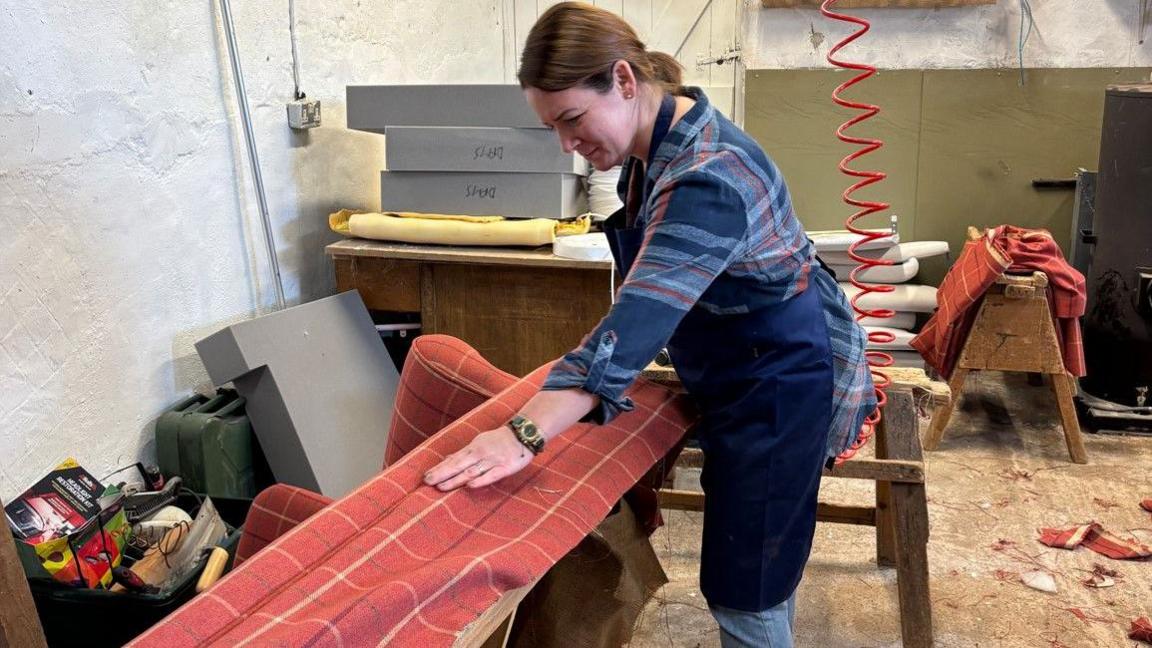 A woman with tied-back brown hair works on reupholstering a sofa lying on its back, stretching patterned red fabric over the frame, surrounded by tools in a workshop.