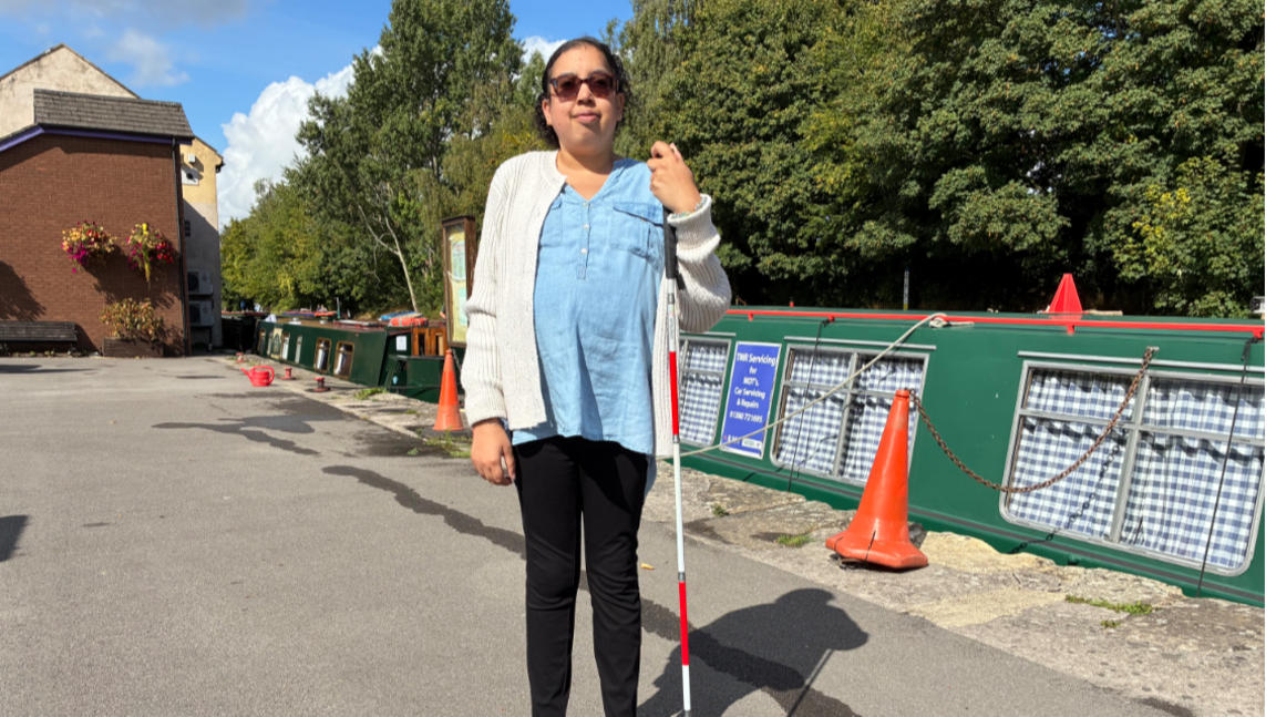 Renee standing by a canal with a cane. She is wearing a blue shirt with a white cardigan and black jeans.