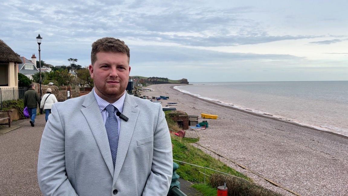 Councillor Henry Riddell wearing a striped shirt and a grey blazer on the seafront in Budleigh Salterton 