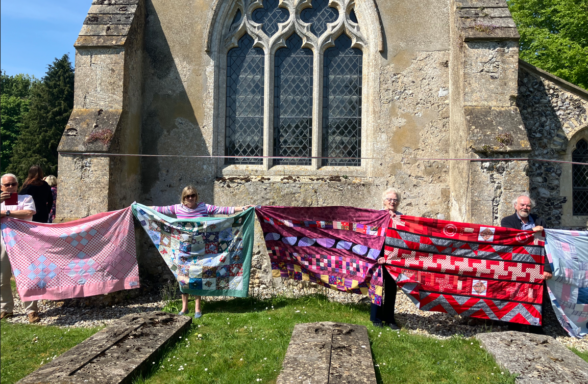 Line of people holding up quilts, with church behind them 