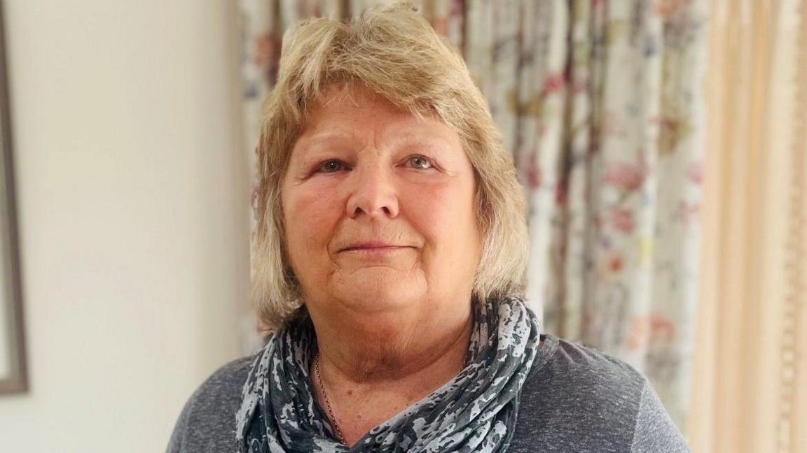 Sandra Geddes, woman with blonde hair and wearing grey scarf and grey top, smiling at camera