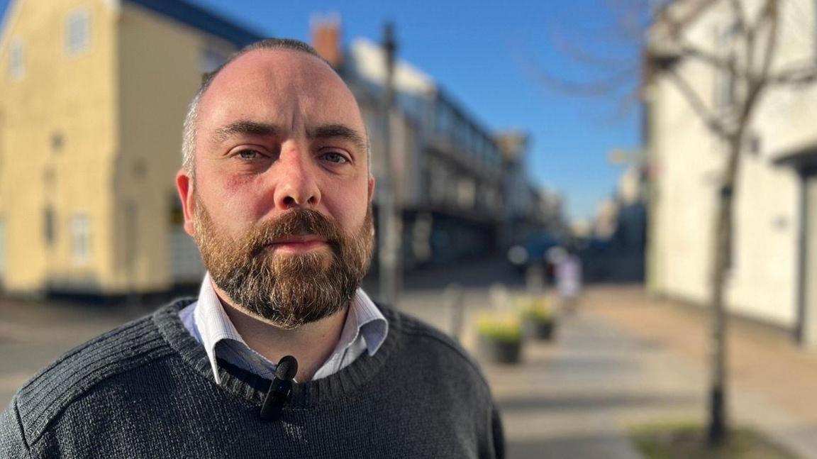 Jake McLean from the GMB union wearing a light-coloured shirt and a dark sweater by the side of the road in Cullompton 