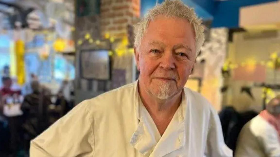 Head-and-shoulder shot of a grey-haired man with a grey beard, smiling in a wry manner. He is wearing a chef's jacket. The blurred background shows a pub or restaurant.
