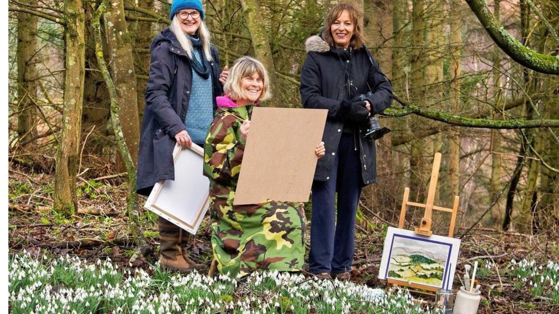 Carey Fluker is in the middle of the picture dressed in a big camouflaged coat. With her are a photographer and another member of the WI. They are in woodland and surrounded by snowdrops.