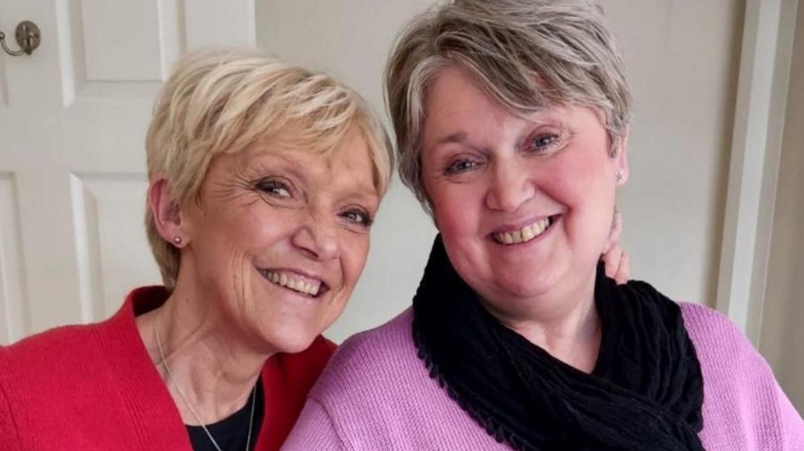 Gillian Wright who is on the left in the photograph and is looking directly at the camera and smiling. She is wearing a black T-shirt with a red cardigan and a silver necklace. Next to her is her sister who is also looking directly at the camera and smiling. She is wearing a pink long sleeve jumper with a black scarf.