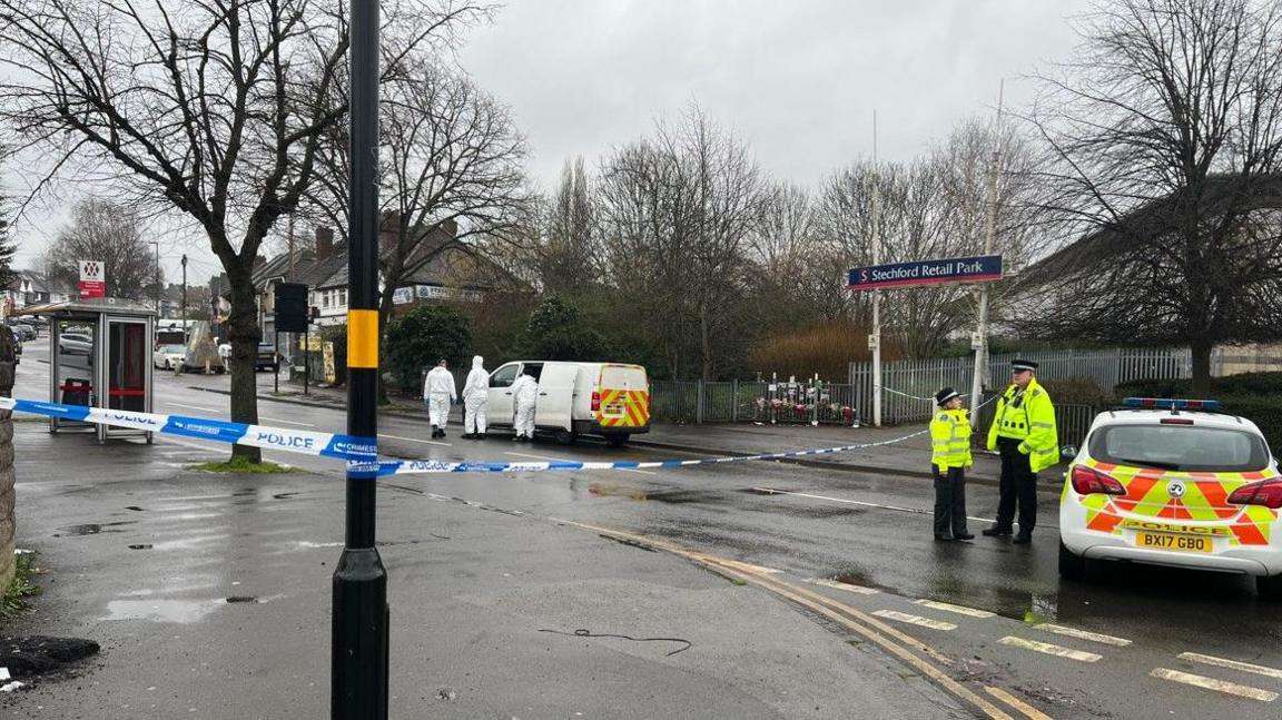 Police tape is wound around a lamp-post on the left hand side of the image and goes across the road on the photo. A police car is on the right  of the photo with two people in police uniform next to it.