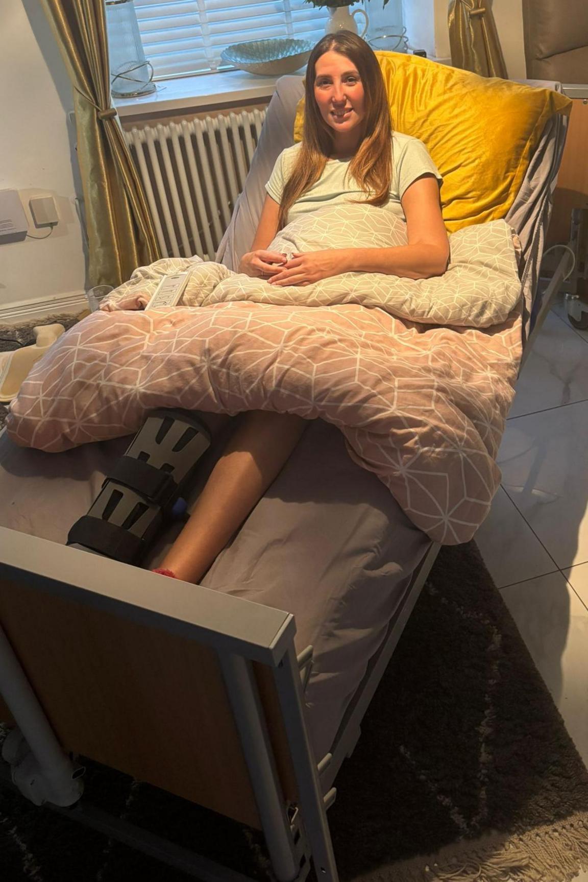 A woman with long brown hair sat in a hospital bed in a living room. One leg is in a boot and she is covered in a pink and grey duvet and propped up on a yellow pillow. Behind her is a window, with curtains and flowers on the windowsill. There is also an armchair in the corner of the room. She is smiling and looking at the camera.