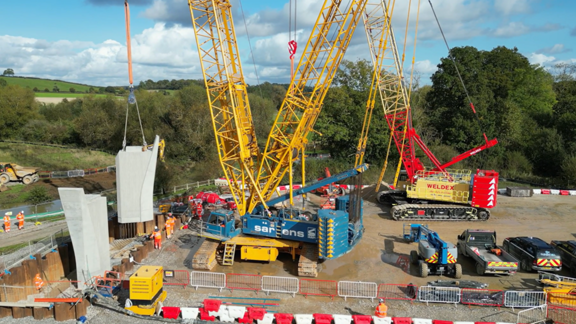 A yellow and blue crane dropping a large concrete block into position alongside another pier that is already in place. There are red and white metal fences around the site and another crane in the background. Empty lorries are parked nearby.  Workers in orange overalls and safety helmets are positioned around the site. There are trees in the background.