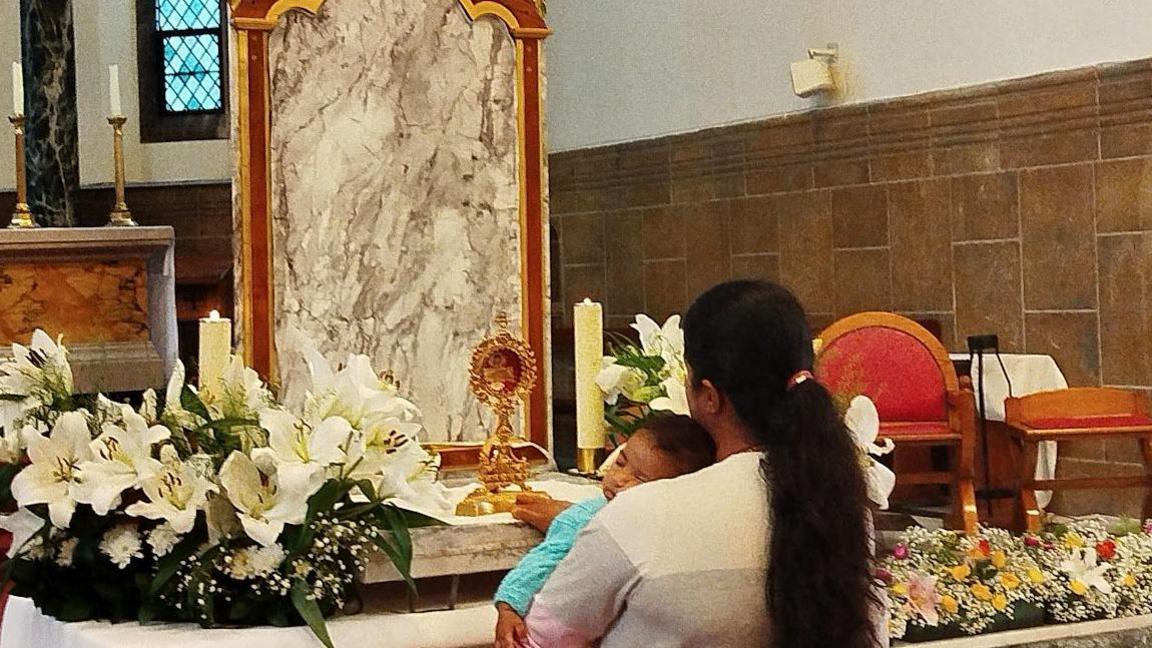 Pilgrim holding a baby prays before the relic of Blessed Carlo Acutis