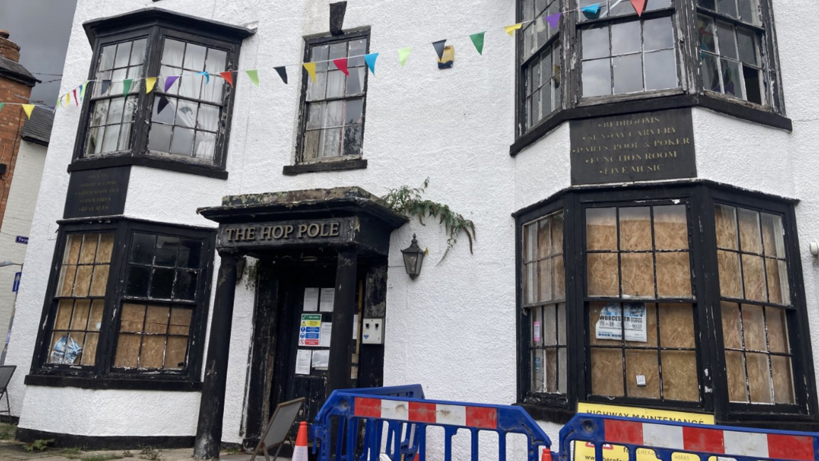 A white three-storey building with peeling black paintwork around the windows and front door, which are boarded up from inside. The letters above the door read The Hop Pole. There is some blue, red and white roadworks fencing and a cone around one of the windows.