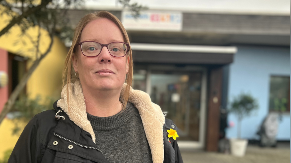 Kerry is wearing a grey jumper, black coat and has a bag over her right shoulder. The centre in the background is painted yellow and red on the left, and blue and yellow on the right. 