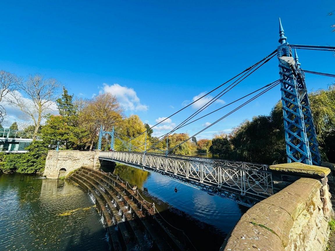 A metal bridge spans a weir with water running under it, between two stone parapets.