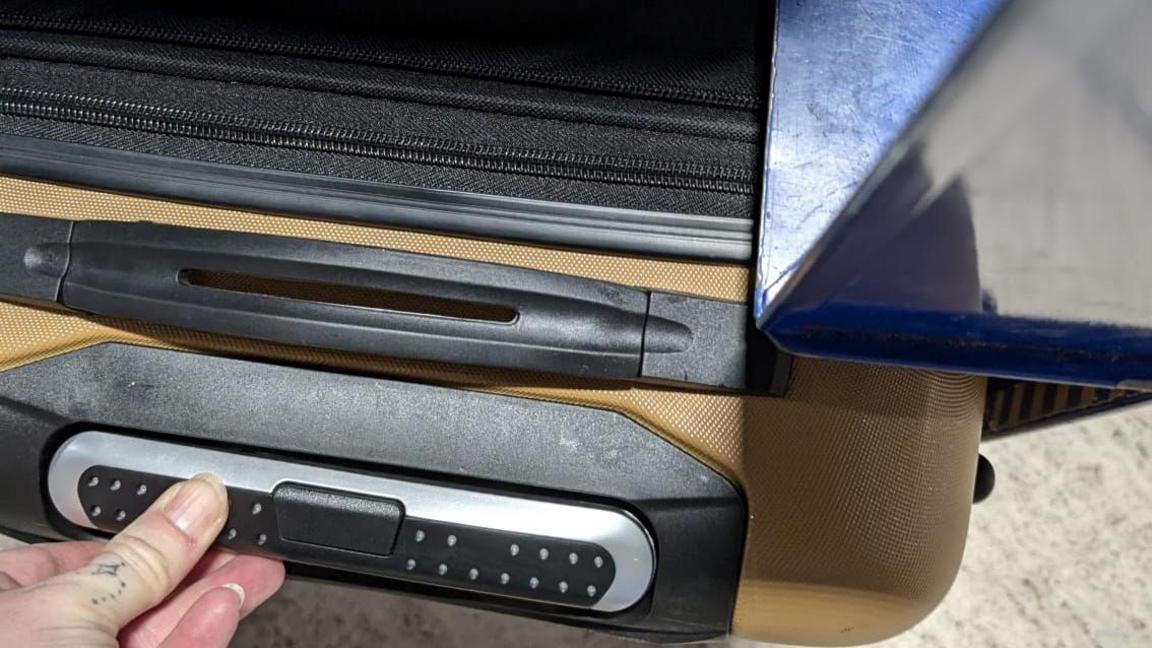 A hand holds a tan suitcase, which appears to fit snugly into a blue box for measuring luggage