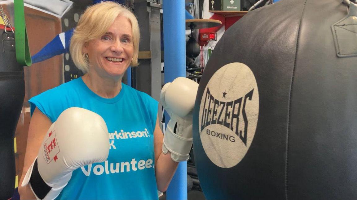 A woman wearing white boxing gloves and a blue t-shirt with the words "Parkinson's UK volunteer" stands next to a punch bag.