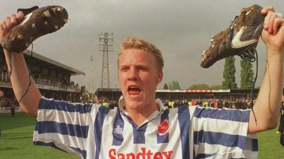Robbie Reinelt lifts up his football boots after scoring an equaliser. He has short blond hair and is wearing a blue and white stripey top.