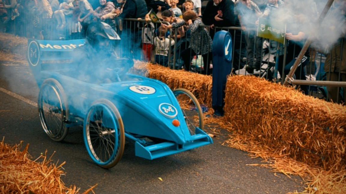 A blue go-kart that is taking part in a race. Crowds stand on either side of the road behind metal fences and hay bales.