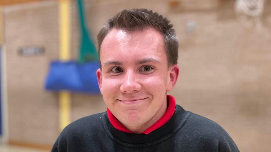 Hamish Crawford smiles at the camera while standing in a sportshall. He has short brown hair and is wearing a black jumper with a red polo top on underneath.