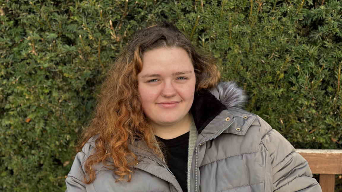 A young woman with long curly hair wearing a grey jacket. She is sitting on a bench and smiling into the camera