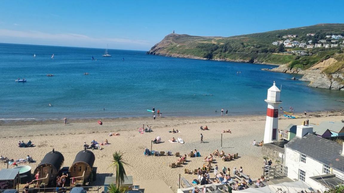 Port Erin in the sun. People sit on a sandy beach by a blue sea. On the right you can see green hills. A white and red lighthouse can also be seen in the forground.