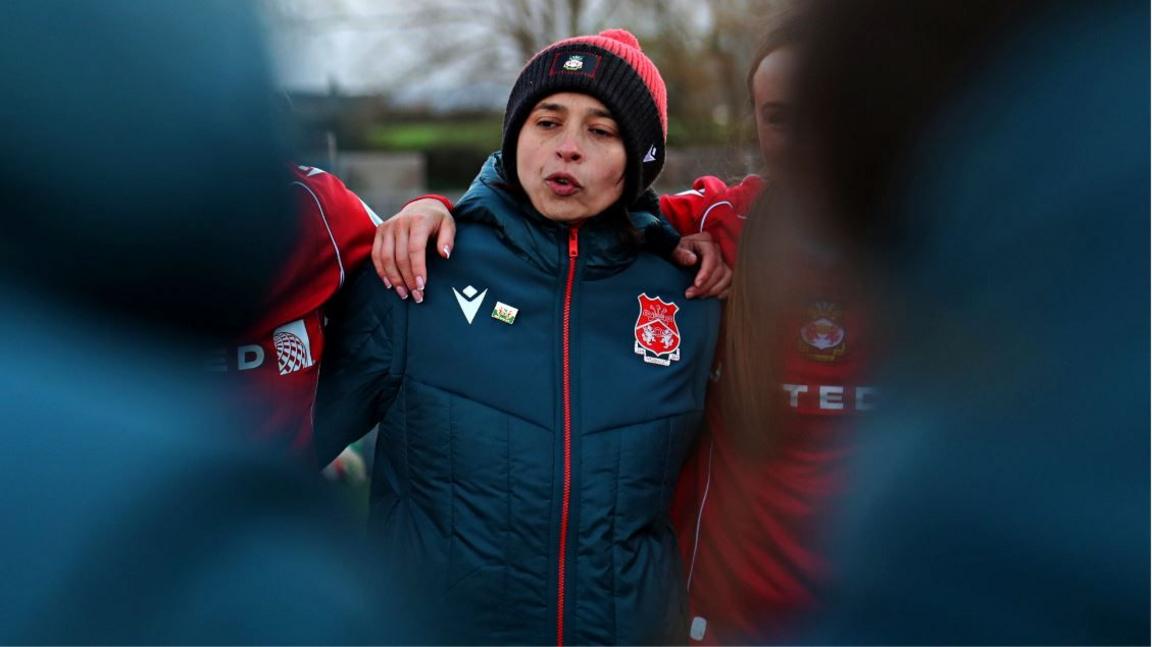 Jenny Sugarman wears a hat and large blue coat as she stands with arms around players in red shirts