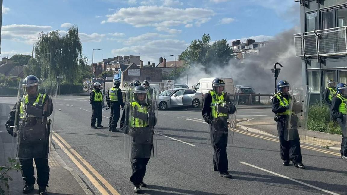 Police officers in riot gear lined across a residential road with smoke emitting from a garage behind