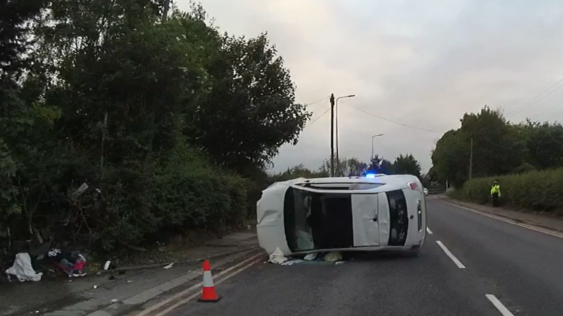 A white Fiat 500 lies on its side in the middle of the road. There is debris on the left-hand side, and a traffic cone in the foreground.