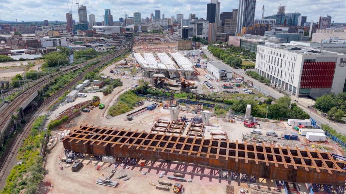 A huge brown bridge structure with a lattice type pattern on the top. It is on a site with vans parked, shipping containers and machinery. On the left is a railway line and in the background are high rise buildings.