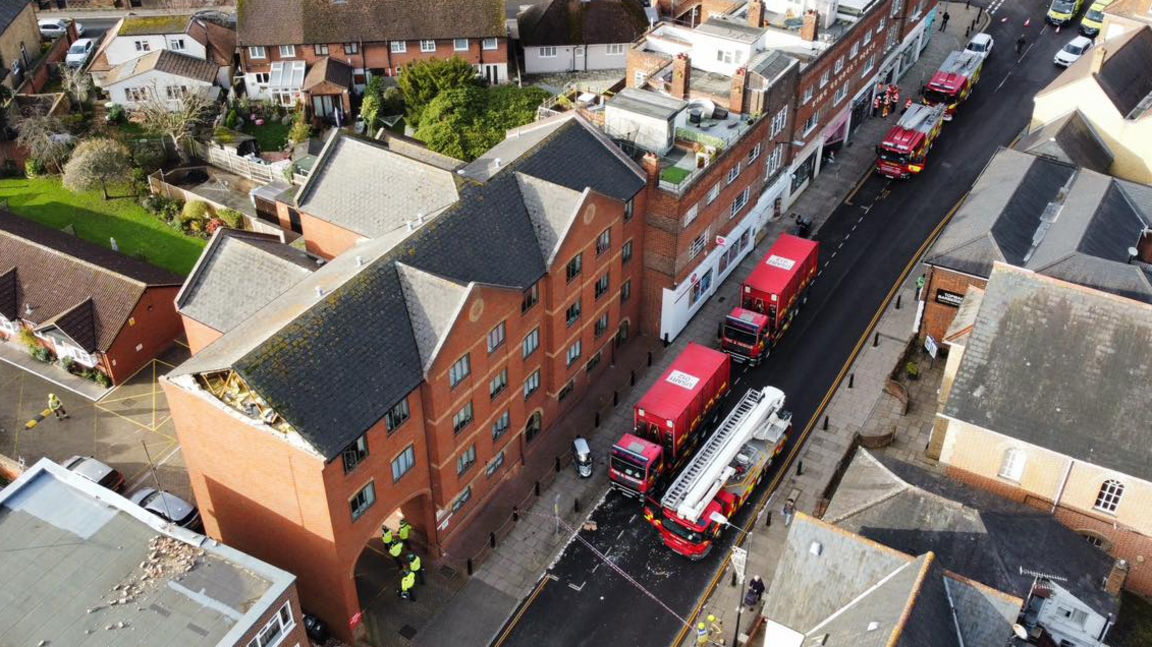 Drone image, taken at an angle, of three fire engines and two red lorries on an otherwise empty High Street. Several fire fighters and police officers are standing around. In the centre of the image a red-brick building has its gable end of bricks missing. Rubble can be seen on a flat roof next door and there is an alleyway between the two buildings. Houses and gardens can be seen behind the buildings.