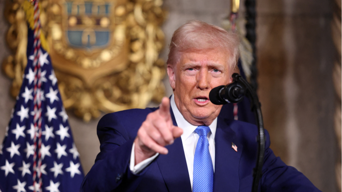 Donald Trump standing at a podium with the US flag visible behind him and a microphone in front of him - he is making a pointing gesture