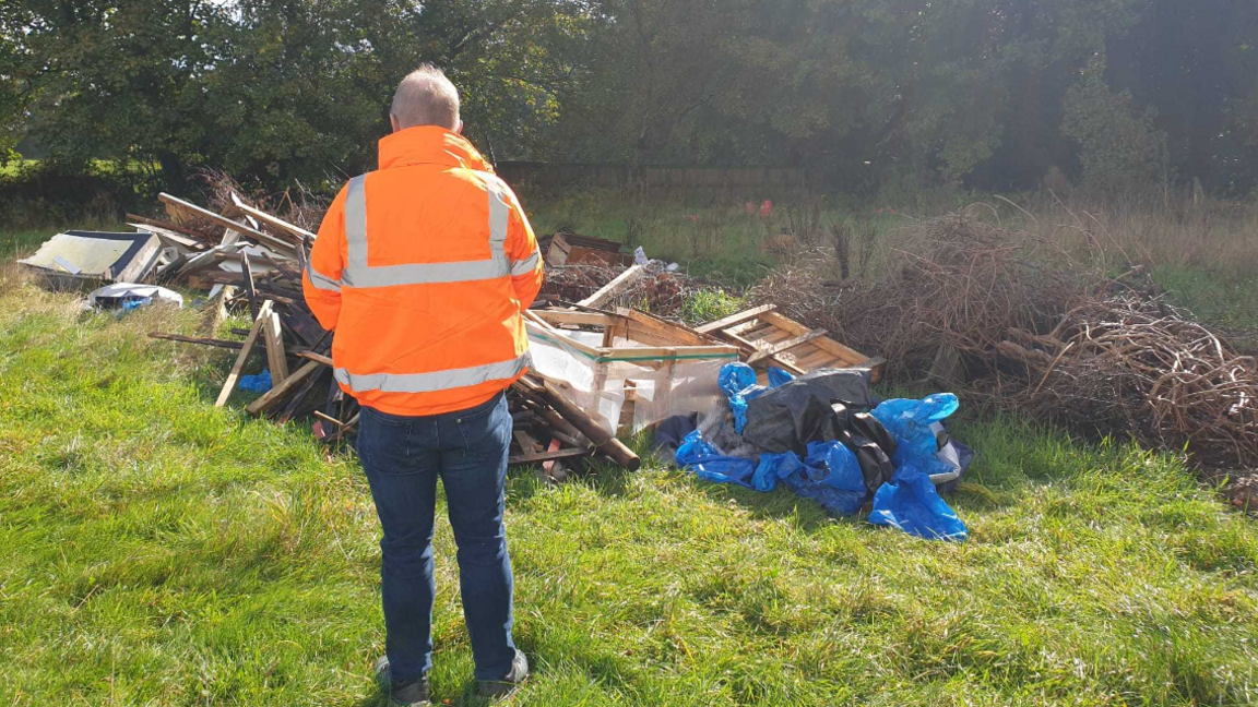 a man in orange high visibility clothing in a field looking at a pile of rubbish