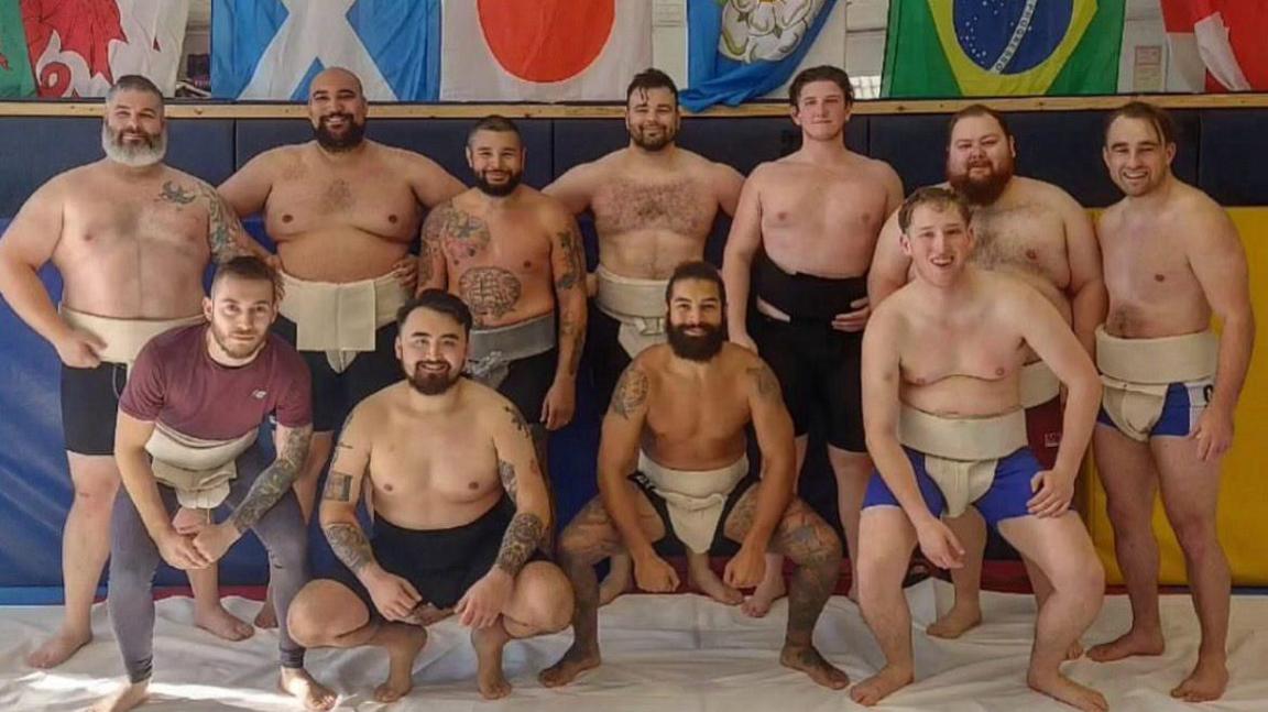 A group of twelve sumo wrestlers pose together on a white mat inside a gym. They wear traditional mawashi belts, with some also in shorts or leggings. Behind them, a wall displays several national flags including Wales, Scotland, Japan, Yorkshire, Brazil, and England. The wrestlers vary in body type and some have visible tattoos. The gym features padded walls and ceiling tiles, indicating an indoor training facility.