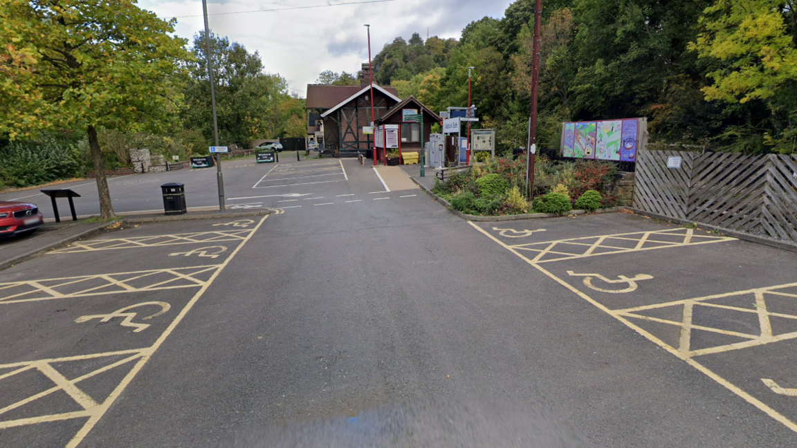 Car park surrounded by trees with a notice board and a small brick station 