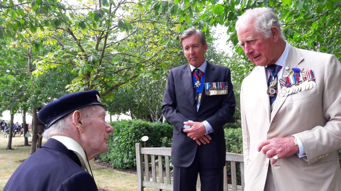 Bill Redston wearing a navy jacket and hat looking up at King Charles wearing a pale suit and medals, with another man in a suit and medals standing with his hands clasped in the background