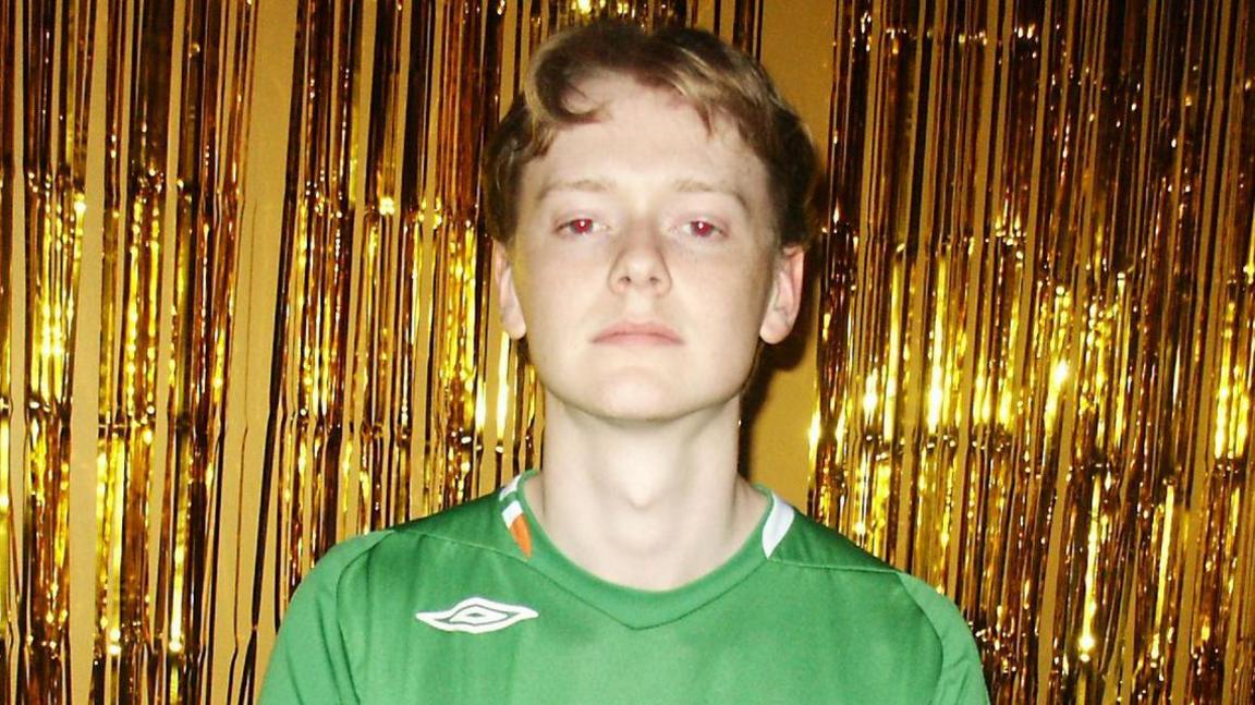 A man with parted red hair is wearing a Republic of Ireland football jersey. The flash from the camera has turned his eyes red. He is standing in front of a long chair that appears to have golden glitter on it. There is also a curtain of golden strips behind him.