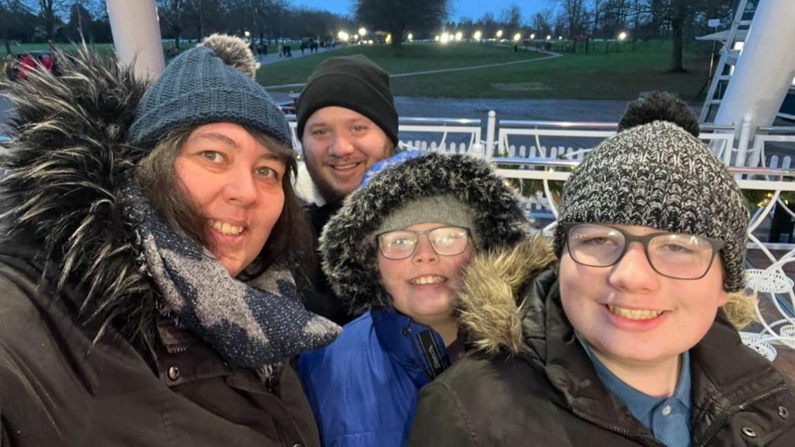 The family smiling for the camera. They are all dressed in warm winter clothes. There is a park behind them and lights could be seen in the background.