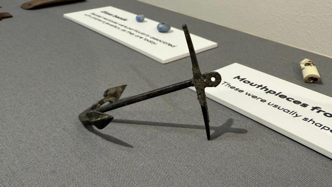 The photo shows a metal anchor which is laid on its side. It has some rust on it. It is laid on a table with a grey cloth. There are artefacts and placards around it. 