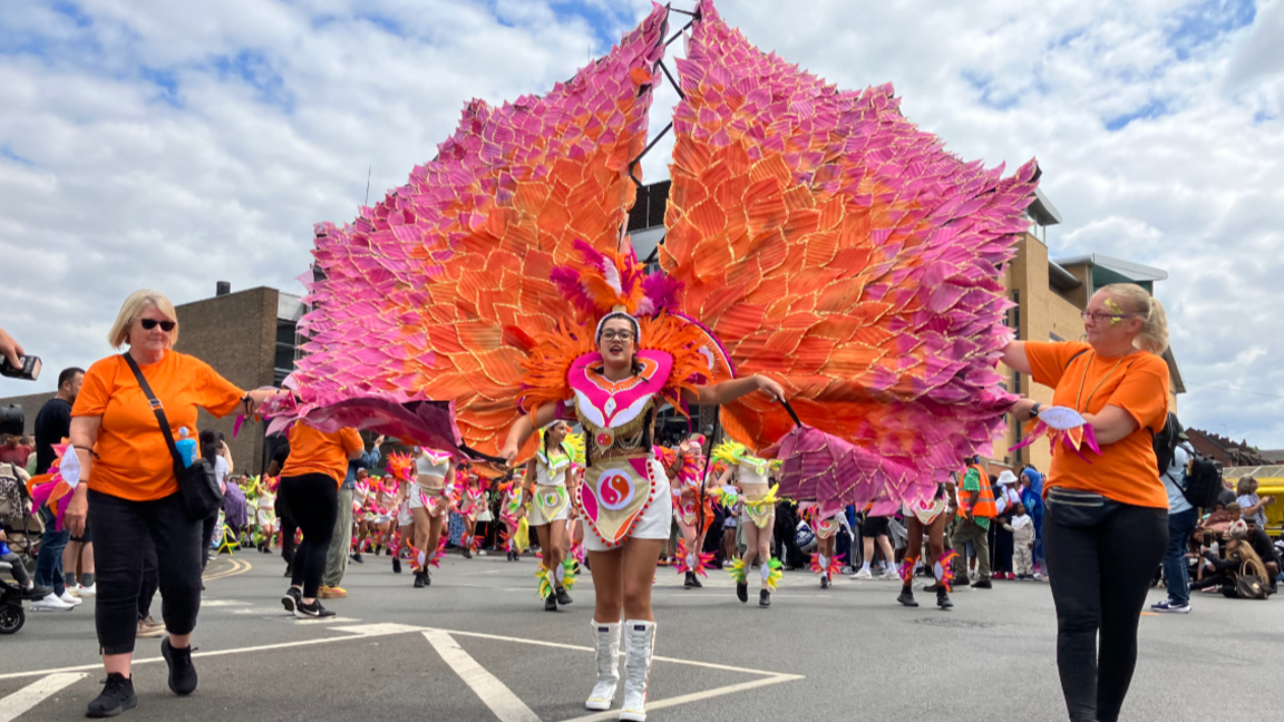 A dancer with huge pink and orange wings attached to her shoulders is parading through Leicester with crowds lining the streets for the city's 40th Caribbean Carnival in 2025.