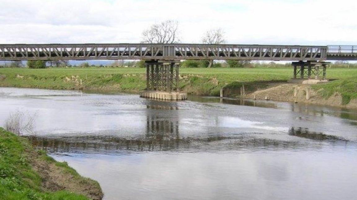 Melverley Bridge, constructed of girders, is supported by struts both over the river and beneath the bridge on land. A couple of trees are in the distance in the middle of the photo.