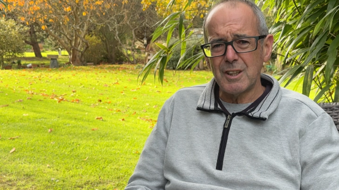 Man with grey top and T shirt and grey hair and glasses with a lawn behind.