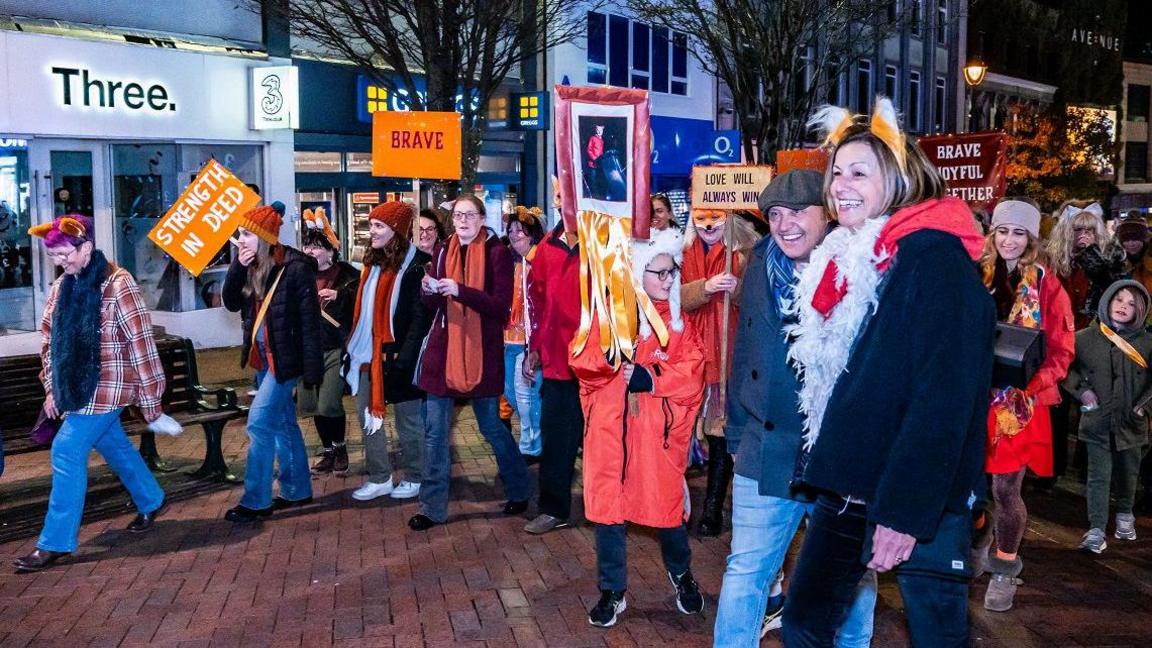 A group of people walking down a high street with shops behind them. They are holding up signs saying "strength in deed" and "brave" and they are all wearing orange clothes and accessories.