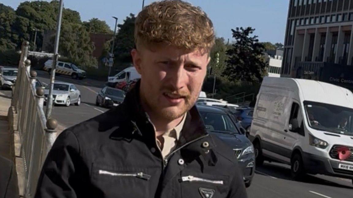 Matthew Stewart leaves court in Carlisle, outside Carlisle Magistrate's Court. He has blonde/strawberry blonde coloured short curly hair and stubble. He is wearing a black biker jacket which is large silver zips. 