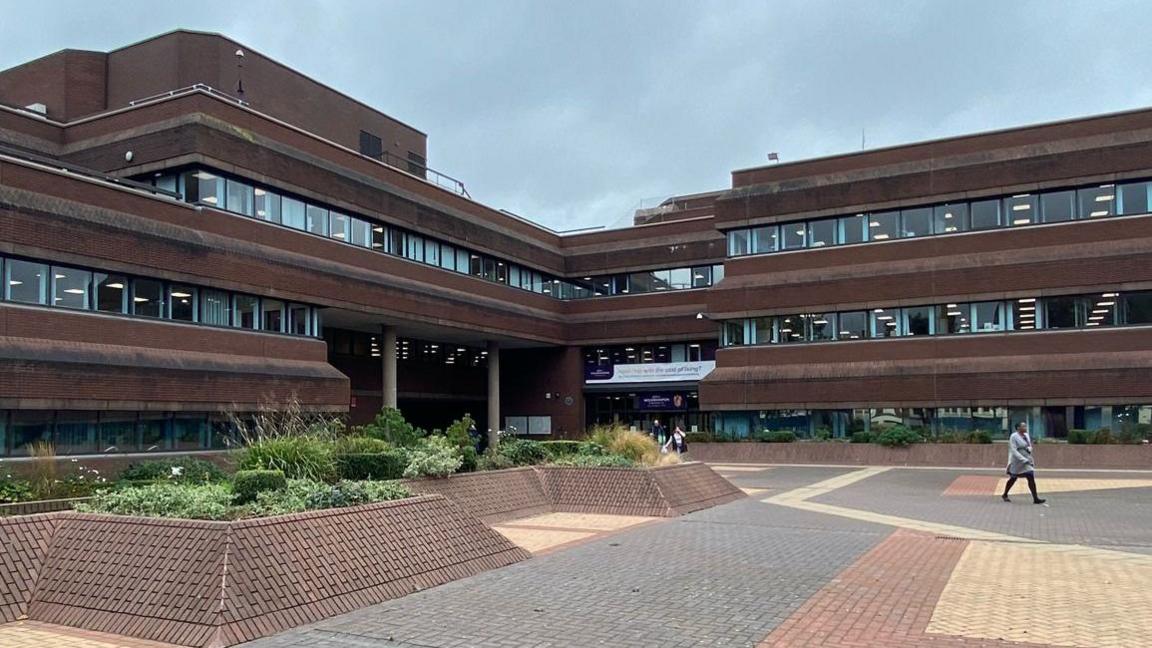 A brown brick brutalist building with green plants outside and a woman in grey walk across the pavement.