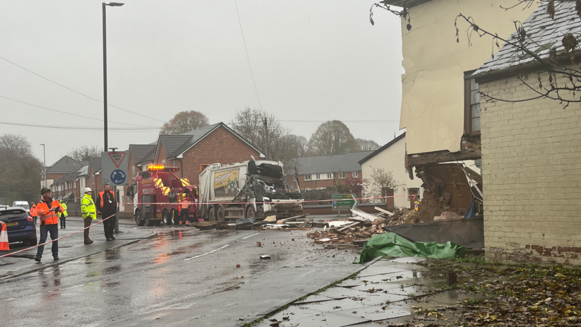 A white bin lorry is towed away from the rubble. The cabin where the crew would have been sat is completely crushed. 