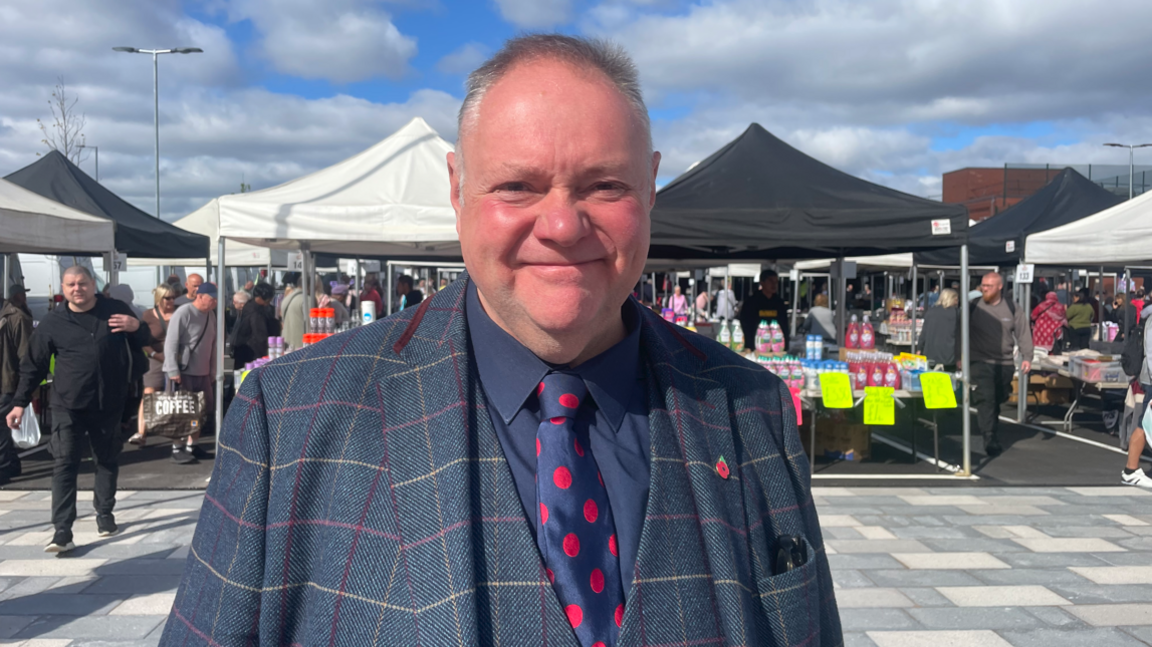 A man wearing a mainly blue jacket and waistcoat, a blue shirt and a blue tie with red circles is looking at the camera.